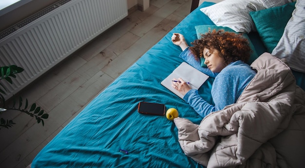 La vista superior de una mujer rizada se quedó dormida en la cama mientras hacía la tarea con un cuaderno cerca y aguantando...