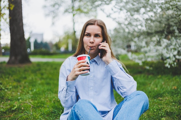 Vista superior de la mujer sentada en el jardín del parque en la hierba verde con el libro de la tableta de los auriculares del teléfono inteligente y el café en la mano