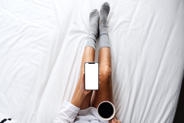 Vista superior de una mujer sosteniendo un teléfono móvil blanco con pantalla de escritorio en blanco mientras bebe café en una acogedora cama blanca en casa por la mañana