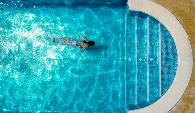 Vista superior de la mujer en traje de baño nadando en agua de piscina transparente con suelo de baldosas azules cerca de las escaleras