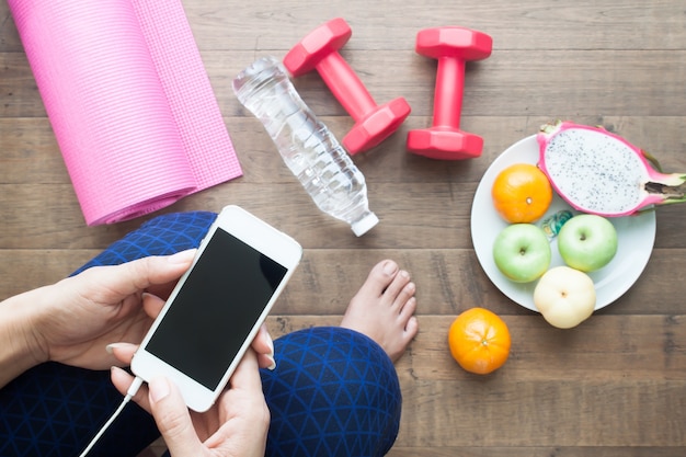 Vista superior de la mujer usando el dispositivo móvil con equipos deportivos y frutas frescas