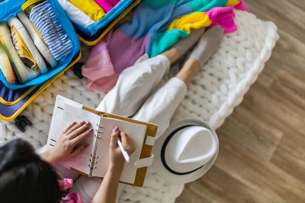 Vista superior mujer viajero tomando lista en bloc de notas en blanco listo vacaciones de viaje de verano sentado en el piso