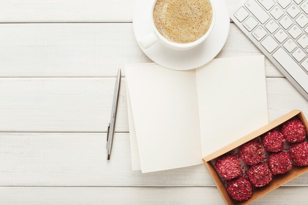 Vista superior na mesa de trabalho com doces vegan crus saudáveis, xícara de café, bloco de notas e teclado do laptop. sobremesa fitness no escritório, copie o espaço