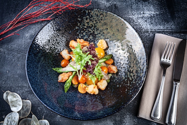 Vista superior na salada quente com salada de camarão, tomate cereja e aspargos servida em uma tigela escura sobre fundo escuro. Comida plana leiga. Feche a vista no almoço com espaço de cópia para o projeto. Frutos do mar