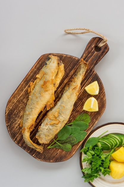 Vista superior de navaga de pescado frito servido en una tabla de cortar de madera con limón, verduras y verduras sobre fondo gris.