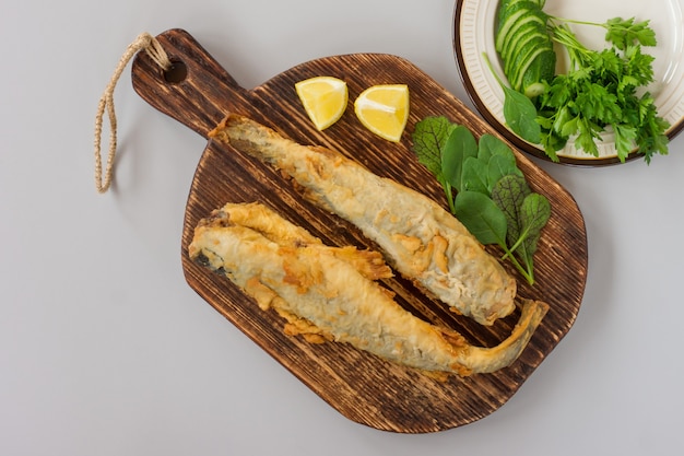 Vista superior de navaga de pescado frito servido en una tabla de cortar de madera con limón, verduras y verduras sobre fondo gris.