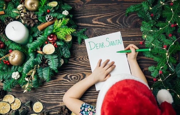 Vista superior niña con una gorra roja escribiendo una carta a Santa Claus con una decoración navideña sobre un fondo. ¡Feliz Navidad y Felices Fiestas!