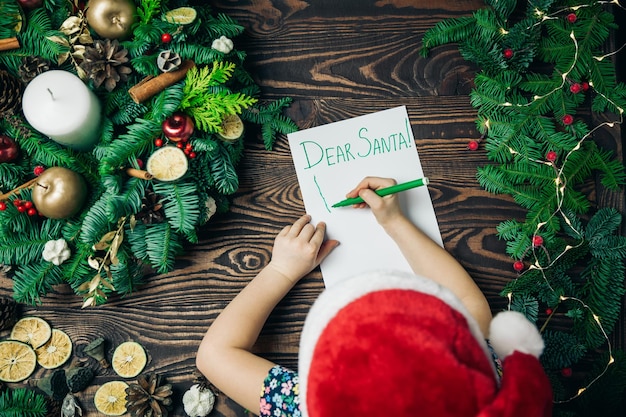 Vista superior niña con una gorra roja escribiendo una carta a Santa Claus con una decoración navideña sobre un fondo
