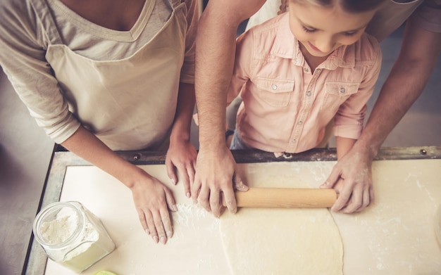 Vista superior de la niña linda y sus hermosos padres aplanando la masa mientras se hornea en la cocina de casa