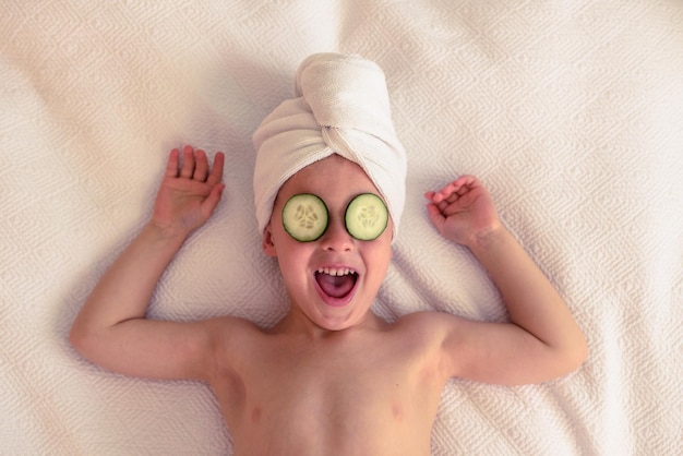 Vista superior de un niño emocionado con una toalla en la cabeza y rebanadas de pepino en los ojos acostado en una manta blanca con los brazos levantados y riendo