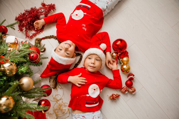 vista superior, un niño y una niña en pijama rojo yacen en el suelo debajo del árbol de Navidad