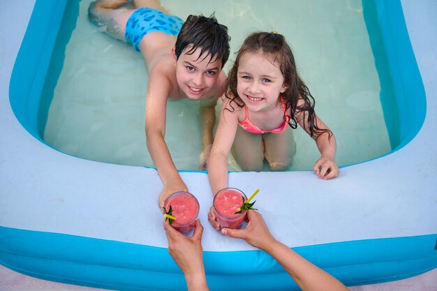 Vista superior de niños lindos en la piscina estirando las manos para tomar una bebida refrescante jugo de fresa recién exprimido