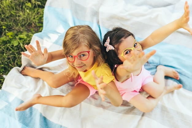 Vista superior de niños positivos con gafas de papel con las manos en alto sentados en la manta Dos niñas pequeñas disfrutando del verano en el parque Amigos divirtiéndose a la luz del sol al aire libre Concepto de infancia