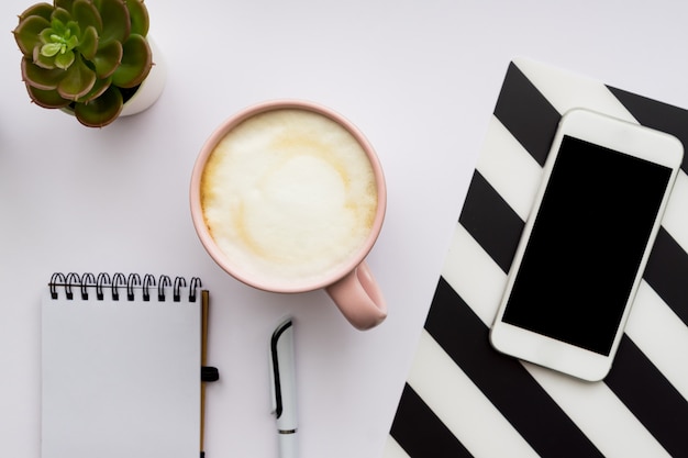 Vista superior de la oficina en casa. Smartphone, bloc de notas vacío y taza de capuchino en la pared blanca