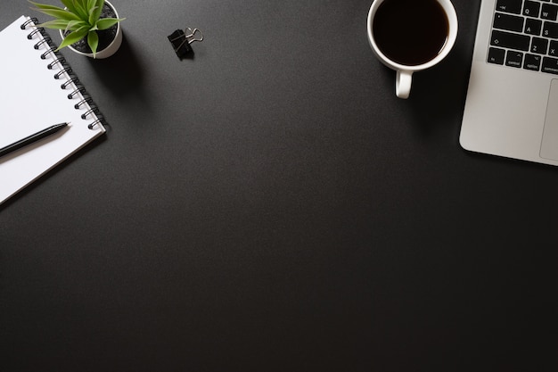 Foto vista superior de la oficina de escritorio con computadora, bloc de notas, bolígrafo, café y planta sobre fondo negro.