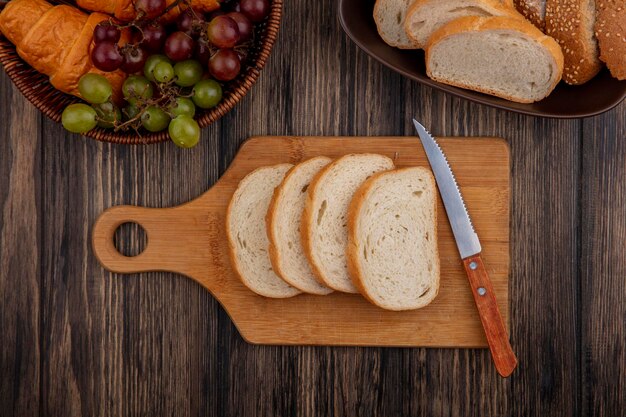 vista superior de panes como rebanadas semillas de maíz marrón y blancos en el cuenco y en la tabla de corte con cuchillo y canasta de uva croissant en fondo de madera