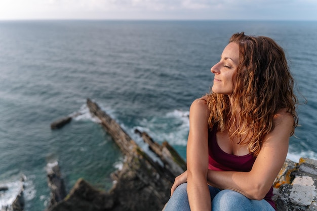 Vista superior panorámica de la mujer con los ojos cerrados de vacaciones en Asturias. Vista horizontal de mujer alegre aislada en el mar azul de fondo. Viajar en vacaciones y concepto de personas.