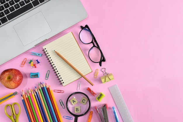 Vista superior de papelería o útiles escolares con libros, lápices de colores, calculadora, computadora portátil, clips y manzana roja sobre fondo rosa.