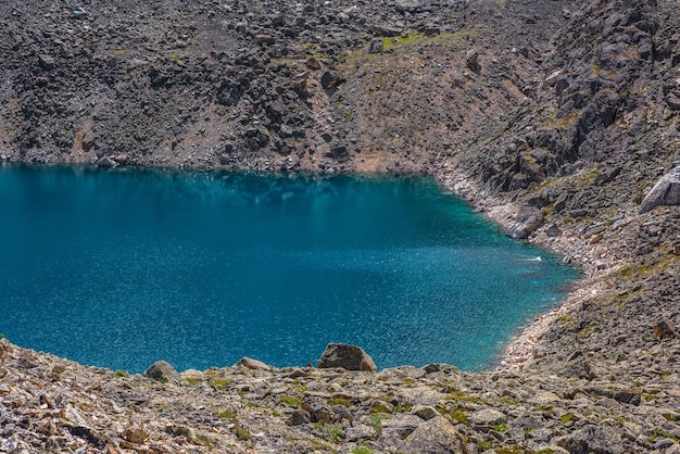 Vista superior para o lago de montanha azul com borda de água ciano brilhante entre rochas iluminadas pelo sol Belo lago alpino com fundo pedregoso em água transparente sob o sol brilhante Fundo natural do lago azul à luz do sol