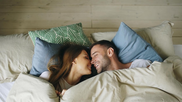 Vista superior de la pareja divirtiéndose en la cama acostada bajo una manta mirando a la cámara y besándose en casa