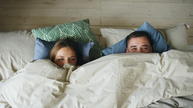 Vista superior de una pareja sonriente divirtiéndose en la cama escondida debajo de una manta y mirando a la cámara