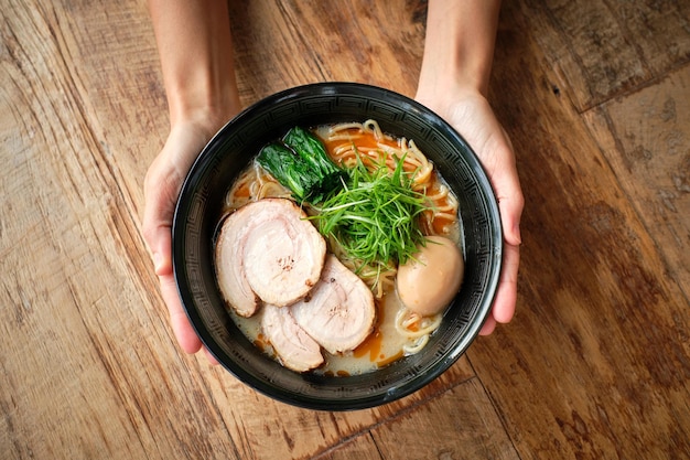 Vista superior de la persona anónima de la cosecha que sostiene un plato negro con sopa japonesa recién cocinada hecha de cerdo cremoso y fideos en una mesa de madera