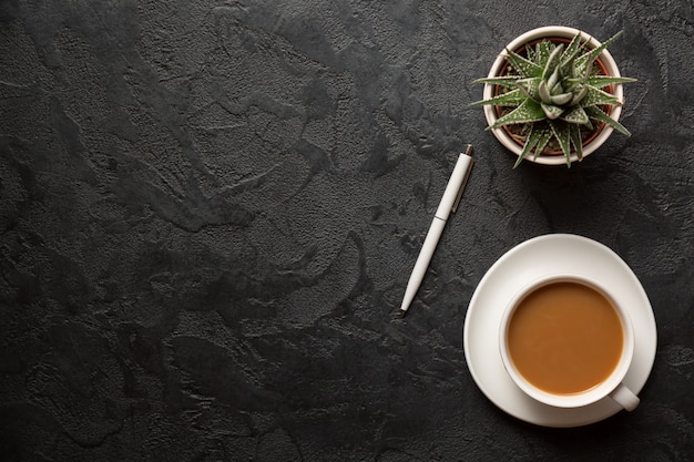 Vista superior plana leigos tiro da mesa da mesa de escritório. Xícara de café cappucino, vaso e caneta em fundo escuro.