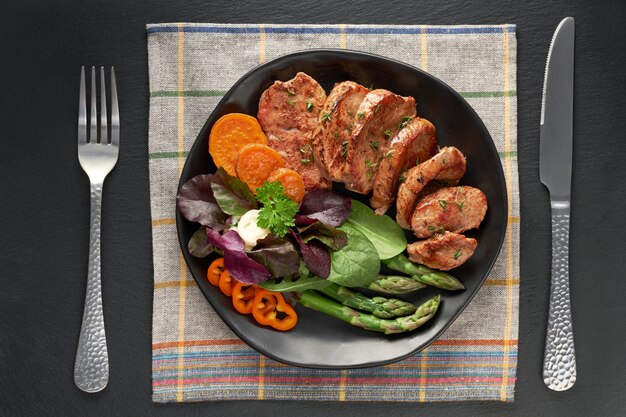 Vista superior de un plato negro con rodajas de carne frita, camote y ensalada mixta en piedra pizarra negra