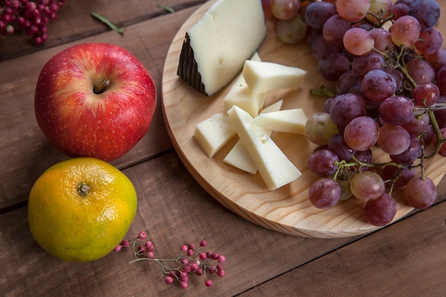 Foto vista superior del plato de queso con uvas, mandarina y manzana roja, sobre mesa de madera