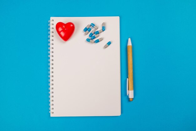 Vista superior de la pluma de la libreta en blanco corazón rojo y pastillas sobre fondo azul aislado