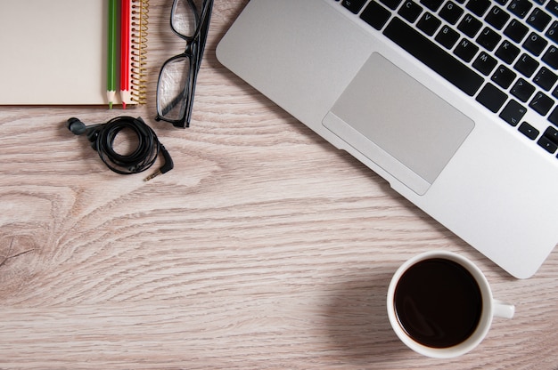 Vista superior del portátil y el bloc de notas con gafas y auriculares en escritorio de madera rústica y taza de café.