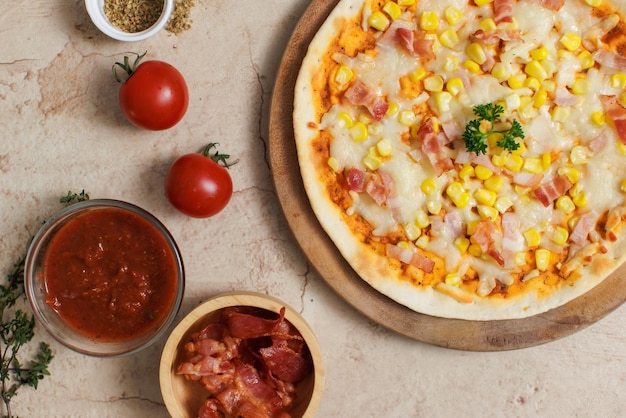 Foto vista superior de primer plano de deliciosa, sabrosa, jugosa, delgada, crujiente, con queso, maíz y jamón, pizza italiana colocada sobre una tabla de madera en una mesa de fiesta alrededor con otros ingredientes, salsa, aceite de oliva, tocino y tomates.