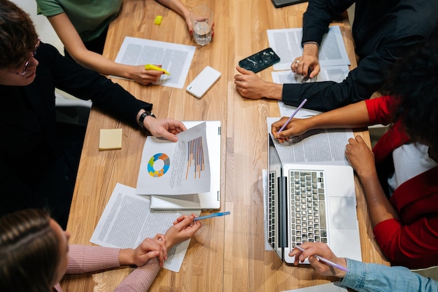 Vista superior de primer plano del equipo de negocios multiétnico que se encuentra en una reunión con colegas inversores actuales discutiendo los datos del gráfico financiero del nuevo plan en la mesa de la oficina con una computadora portátil