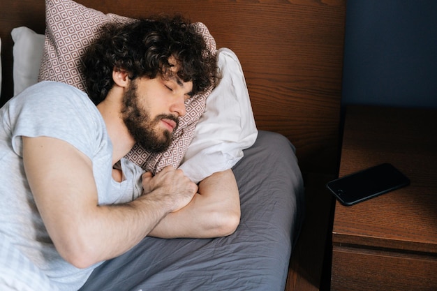 Vista superior de primer plano de un joven barbudo caucásico durmiendo pacíficamente acostado de lado en una cama doble grande y cómoda bajo una manta blanca