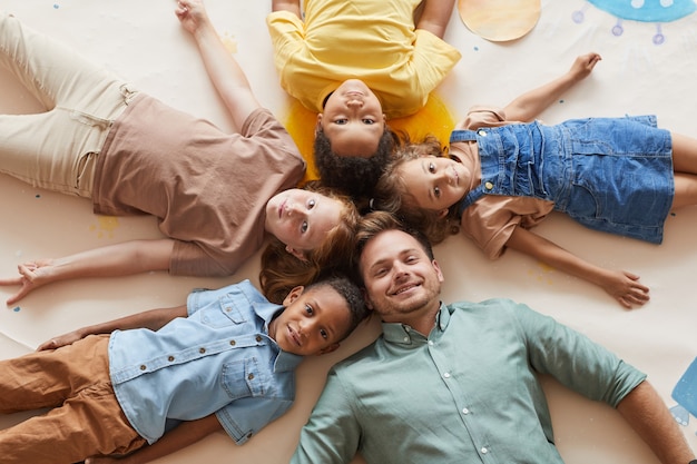 Vista superior del profesor sonriente con un grupo multiétnico de niños que yacen en círculo mientras se divierten en el centro de preescolar o desarrollo.