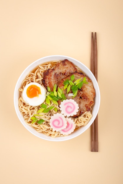 Vista superior del ramen de sopa de fideos japoneses en un tazón de cerámica blanco con fideos, caldo de carne, cerdo asado en rodajas, cebolla verde, narutomaki, yema de huevo, fondo beige. Plato tradicional de Japón, primer plano