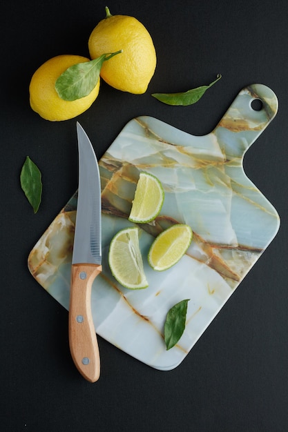 Vista superior de rodajas de limón y cuchillo en tabla de cortar con limones y hojas sobre fondo negro