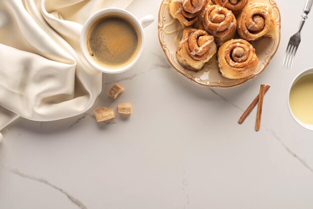 Vista superior de rollos de canela caseros frescos en la superficie de mármol con una taza de café con azúcar moreno