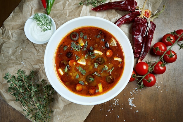 Vista superior de la sabrosa sopa rusa espesa, picante y amarga de Solyanka con aceitunas, limón y salchichas en un tazón blanco sobre mesa de madera.