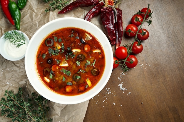 Vista superior de la sabrosa sopa rusa espesa, picante y amarga de Solyanka con aceitunas, limón y salchichas en un tazón blanco sobre superficie de madera.