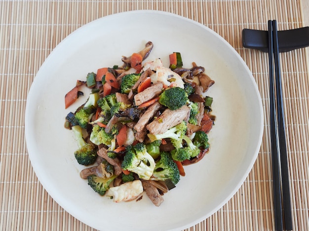 Vista superior de salteados en un plato con verduras, proas y carne. palillos negros sobre un mantel de bambú