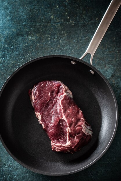 Vista superior de una sartén oscura con un trozo de carne cruda sobre una superficie de mármol.