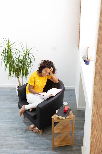 Vista superior de la sonriente mujer morena leyendo un libro en casa. Espacio para texto.