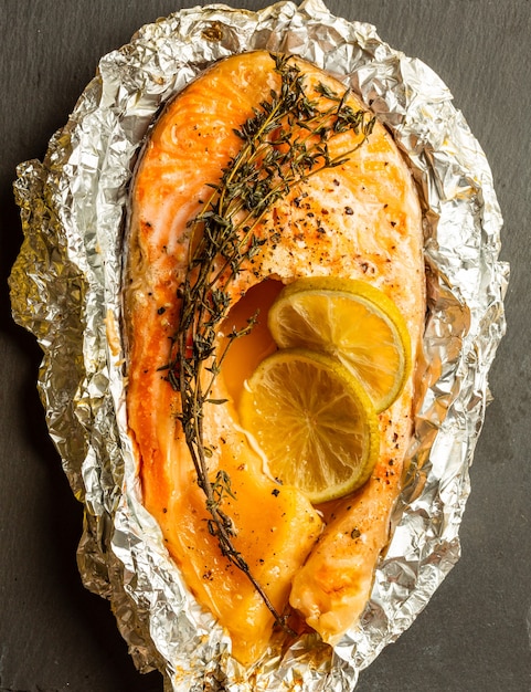 Vista superior de la superficie de pizarra negra con sabroso pescado al horno en papel de aluminio con rama de tomillo y rodaja de limón