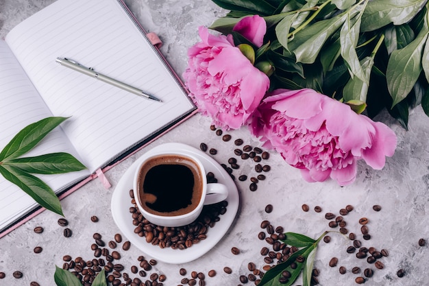 Vista superior de la tarjeta de café en una taza junto a peonías y un cuaderno sobre un hormigón gris brillante