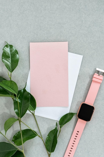 Vista superior de la tarjeta rosa vacía con reloj de sobre blanco y hojas sobre fondo gris