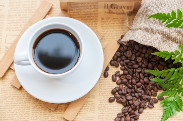 Vista superior de la taza blanca de café negro en la placa de madera sobre el grano de café borroso