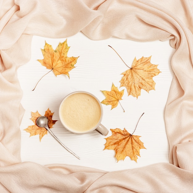 Vista superior de la taza de café capuchino con espuma de leche, bufanda de tela cálida y hojas amarillas de temporada otoñal de arce en la mesa de madera blanca