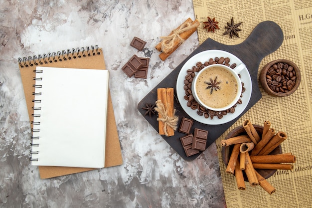 Vista superior de una taza de café con galletas de anís granos de café tostados en un platillo chocolate palitos de canela en un tazón anís en un tablero de madera cuadernos en la mesa