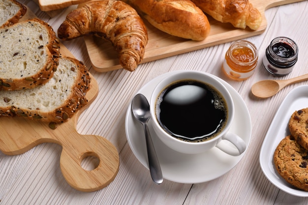 Vista superior de la taza de café con pan o bollo, croissant y panadería en mesa de madera blanca